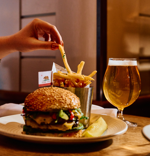 Hand holding a refreshing drink with a cucumber garnish against a green-tiled background.