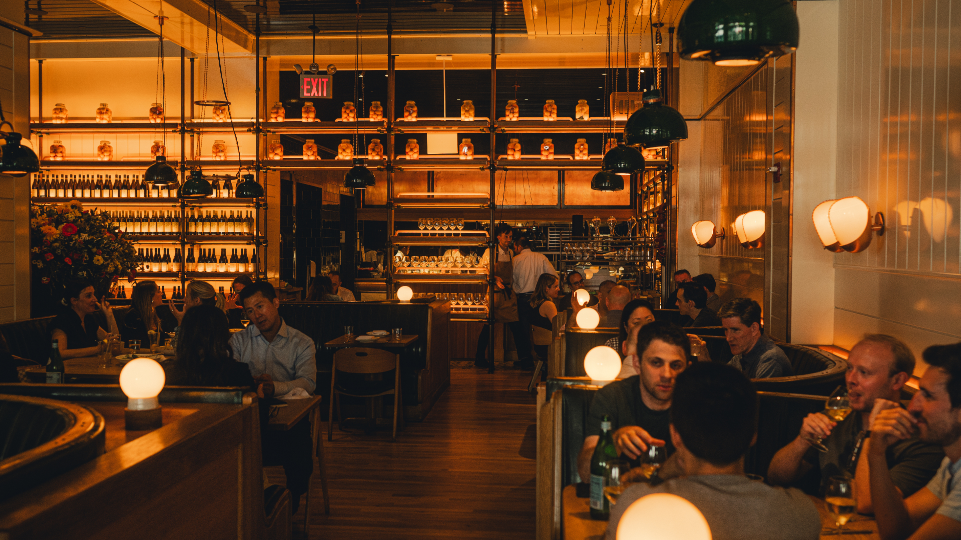 A cozy restaurant interior with wooden tables, neatly set for dining, and a bar with green-tiled walls and a bartender preparing drinks.