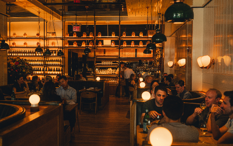 Modern restaurant interior with wooden tables, bar seating, pendant lighting, and cream-colored walls with vertical paneling