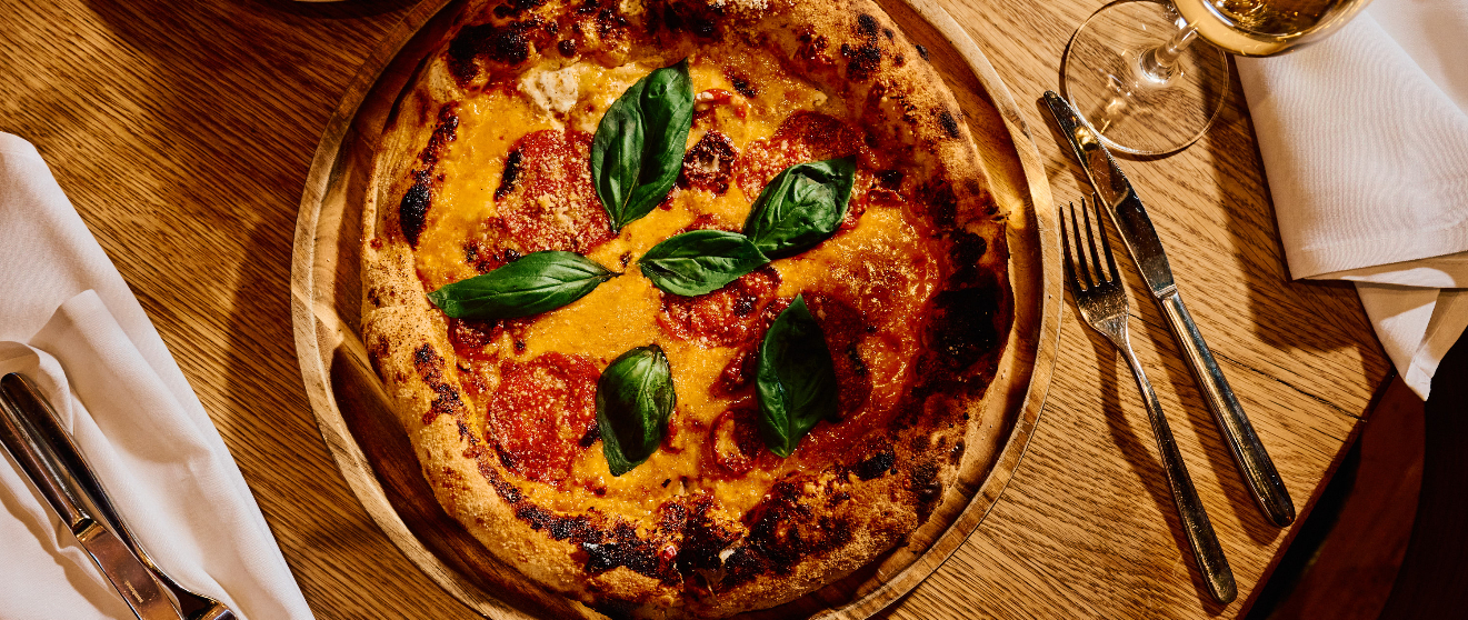 Wood-fired pizza with tomato sauce, melted cheese, and fresh basil leaves on a wooden board with wine and cutlery