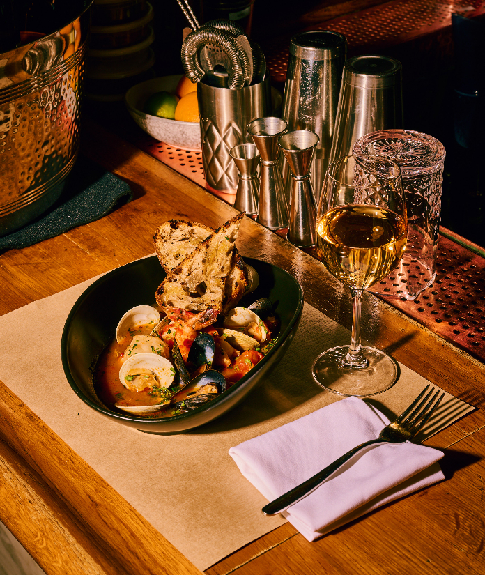 Seafood dish with clams and mussels in black bowl served with bread and white wine on wooden table with bar tools