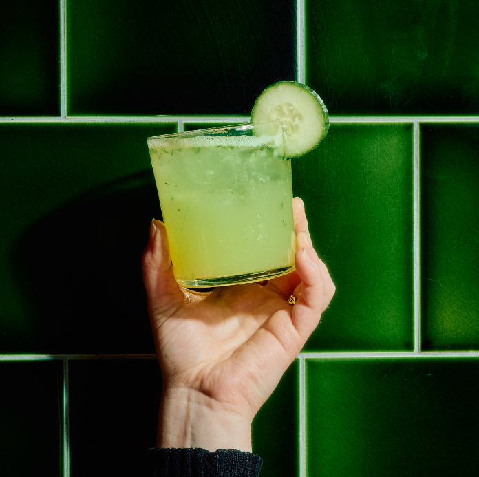 Hand holding a pale green cocktail with cucumber garnish and ice against green tile background