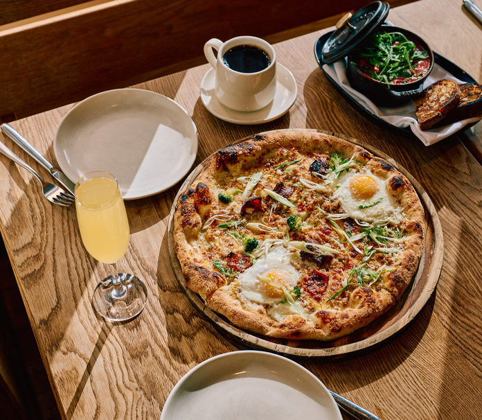 Wooden table with cooked pizza topped with eggs and greens, a glass of juice, a cup of coffee, and a small bowl of salad.