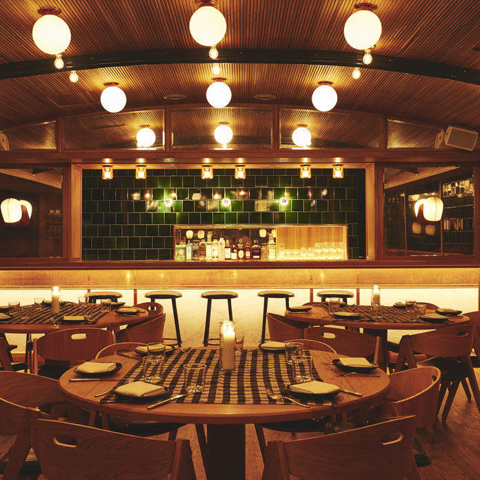 Modern restaurant interior with round wooden tables, burgundy chairs, globe lighting, and green tile bar backdrop